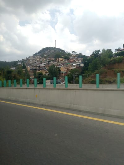 The image depicts a view from a highway overlooking a densely populated hillside in Abbottabad, Pakistan. The foreground features a concrete barrier with several green cylindrical posts, separating the highway from the embankment. A yellow line marks the edge of the road. The midground shows a hillside densely packed with houses and buildings of varying colors and sizes, many built on steep slopes. Trees and vegetation are interspersed among the buildings. In the background, the sky is overcast with thick grey clouds, and a tall communication tower is visible on the hilltop. The overall scene suggests a built-up urban area on a mountainous terrain, characteristic of Abbottabad. There are no people visible in the image.