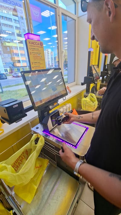 A medium shot captures a person at a self-checkout kiosk in a retail store, likely in Dzyarzhynsk, Belarus, as suggested by context. The individual, viewed from the chest up and primarily in profile, wears a black t-shirt and sunglasses. Their right hand holds a black object, possibly a bottle, poised above the scanning platform. A silver watch is visible on their left wrist. The kiosk features a prominent touchscreen display showing product images, a scanner, a scale with purple LED lighting, and a receipt printer. Yellow plastic shopping bags are present on the scale and in a basket to the left. Behind the kiosk, a window reveals a view of apartment buildings and cars on a street, consistent with an urban setting. The store interior is painted yellow and white, with fluorescent lighting overhead.