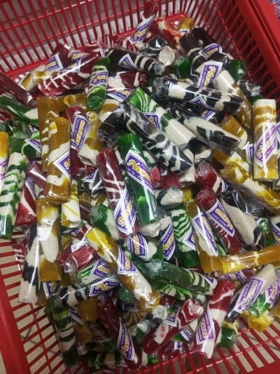 The image displays a large quantity of individually wrapped, multi-colored dodol candies, identified by visible labels as "RADJA DODOL," contained within a red slatted plastic basket. Each candy is cylindrical or irregularly shaped, approximately 10-15 cm long, and encased in clear plastic film. The sweets exhibit a variety of color combinations, often marbled or striped, with prominent hues of green, red, yellow, dark brown, black, and white/cream. Many wrappers feature a purple and white label with "RADJA DODOL" and a product registration number. The candies fill the basket to the top. No individuals are present, and no actions or interactions are depicted. The environment suggests an indoor retail or storage setting within Lê Thanh Nghị Ward, Vietnam.