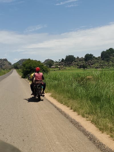 An overhead, outdoor, medium shot captures a rural road in Panyam, Nigeria, under a bright, partly cloudy sky. Two individuals are riding on a motorcycle, moving away from the camera along a paved road. The passenger in the back wears a pink shirt and a red headscarf, while the driver wears a yellow shirt. The motorcycle appears to be dark-colored. Tall, green grass lines the right side of the road, with a dirt and gravel shoulder. Beyond the grass, a field of crops stretches towards a rocky, tree-covered hillside in the distance. The landscape suggests a dry, warm climate.