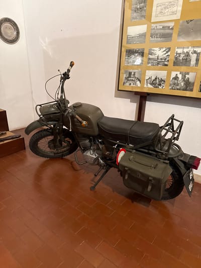 A military-green motorcycle is centrally displayed indoors at a location in Praia do Ribatejo, Portugal. The vehicle features a black single seat, knobby tires, an olive-drab fuel tank, and high handlebars with orange turn signals. It rests on a kickstand. A military-green canvas-like saddlebag is affixed to the right rear, with a red and white circular reflector mounted above it. A partial license plate, reading "B 0U6," is visible on the rear fender. The floor is covered with reddish-brown rectangular tiles. The walls are white. On the left wall, a circular metallic decorative plate is mounted. Below it, a dark wooden multi-tiered stand displays an elongated dark object. On the right wall, a large framed exhibit contains a grid of approximately 15 black-and-white photographs and documents.