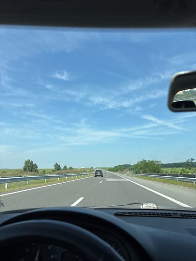The image depicts a forward-facing perspective from within a vehicle traveling on a multi-lane highway near Krumovo Gradishte, Bulgaria, under a bright blue sky with scattered cirrus clouds.
The upper frame is dominated by the clear sky. The distant horizon reveals expansive green fields on the left and forested, rolling terrain on the right. A red and white "GAS" sign is faintly reflected in the car's rearview mirror, which is visible in the upper right quadrant.
The midground features a dark asphalt highway with distinct white dashed lane markings and solid white shoulder lines. Metal guardrails, white and silver, flank both sides of the road, which is bordered by green vegetation and deciduous trees. Two dark sedans are visible ahead in the same direction of travel, receding into the distance; one in the right lane is more distinct than the other, further ahead in the left lane.
The immediate foreground shows the vehicle's interior: a dark dashboard with air vents, and the upper section of a black multi-spoke steering wheel. Through the steering wheel, a speedometer dial on the instrument cluster is partially visible, along with an illuminated red warning light on its lower right section. No people are visible.