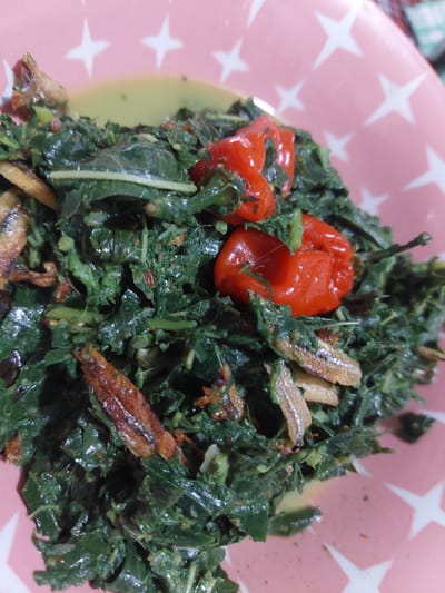 A close-up, overhead shot shows a pink plate with white stars, filled with a dish of cooked green leafy vegetables and small, fried fish. Two whole red chilies are visible in the center of the dish. The food appears to be in a light sauce. The lighting suggests an indoor setting. The dish is likely a local Indonesian cuisine, possibly from the Jelutung area in Indonesia.