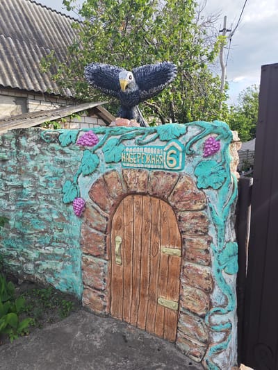 This is a medium shot, eye-level, outdoor, horizontal photograph. The image features a decorative garden wall with a faux door and an eagle sculpture. The wall is painted in shades of turquoise and teal, with a textured, weathered appearance. It is adorned with sculpted grapevines and clusters of purple grapes. The faux door is made to look like wood with visible grain and has brass-colored hinges and a handle. Above the door, a light blue sign with raised yellow lettering reads "НАБЕРЕЖНАЯ 6," which translates to "Embankment 6." Atop the wall, a black and white eagle sculpture with outstretched wings and a yellow beak is positioned. In the background, a grey, corrugated roof of a building is visible, along with green trees and a power pole. The sky is partly cloudy, suggesting it might be daytime. The overall scene appears to be in a residential garden or yard in Krutinskiy, Russia.