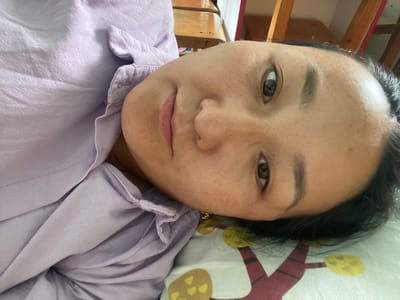 The image is a close-up, sideways shot of a person's face and upper body, lying down. The person appears to be an adult with fair skin, dark hair, and brown eyes. They are wearing a light purple, collared shirt. The background includes furniture, possibly a desk and chairs, suggesting an indoor setting. The overall lighting is soft. It is not possible to determine the location within the city of Киров, Kyrgyzstan from the provided image.
