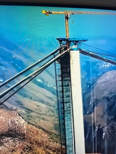 The image shows a close-up of a large cable car structure, possibly under construction or in operation. The main subject is a long, enclosed car with a light-colored exterior, suspended by thick cables. The car has a glass front, offering a view of the interior and potentially the surrounding landscape.
The car is supported by a complex metal framework, painted in dark blue and black. A yellow crane arm extends from a structure on the left side, suggesting ongoing work or maintenance. The cables are a prominent feature, stretching from the car upwards and to the sides, disappearing out of frame.
The background is a mix of sky and a mountainous or hilly terrain. The sky is a clear, bright blue. The ground below appears rocky and sparsely vegetated, with patches of green and brown. The overall impression is one of a vast, open space, possibly a natural landscape. The image is taken from a slightly elevated perspective, looking down at the cable car and the terrain.
There is no visible text in the image. The time of day appears to be daytime, with bright natural light. The weather seems clear and sunny. The scene conveys a sense of scale and engineering, with the large structure contrasted against the natural environment. No people are clearly visible, although there might be some figures within the cable car or on the crane, too small to discern. The activity appears to be either transporting passengers or undergoing construction/maintenance. The specific location context of Vangaži, Latvia, suggests this might be a particular cable car system within that region, possibly for tourism or infrastructure.