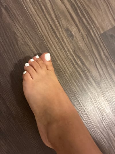 A close-up, overhead shot shows a person's left foot with freshly painted white toenails resting on a dark brown, wood-patterned floor. The foot is positioned from the ankle down, with the toes slightly spread. The lighting is overhead, casting subtle shadows on the floor. The image was taken in Balch Springs, United States of America.