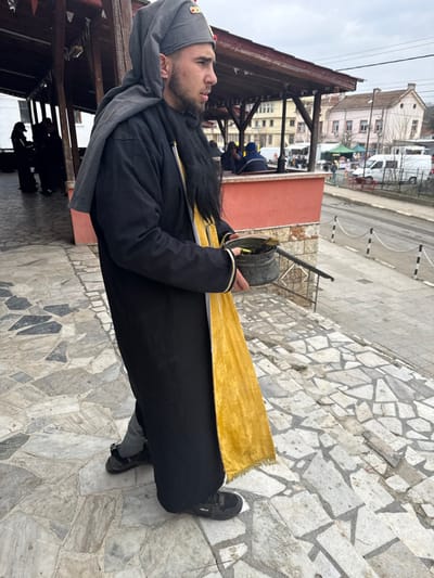A young male is depicted in side profile, facing right, standing on irregular light-colored stone steps in Zidarovo, Bulgaria. He is dressed in a grey conical cap with a reddish-gold emblem, a long black robe, a yellow cloth sash draped over his left shoulder, and a black artificial beard covering his lower face and chest. In his left hand, he holds a dark, weathered metal pot containing dark material, and a thin, dark stick or rod extends downwards from the pot, held by his right hand. He wears black sneakers with white soles. To his immediate left and rear, under a wooden-framed shelter, other blurred individuals are present. To his right, below the steps, a street is visible, bordered by a pavement barrier consisting of metal posts connected by chains. Across the street, multiple light-colored buildings with tiled roofs, parked white vans, and a distant outdoor market with green awnings are visible under an overcast sky.