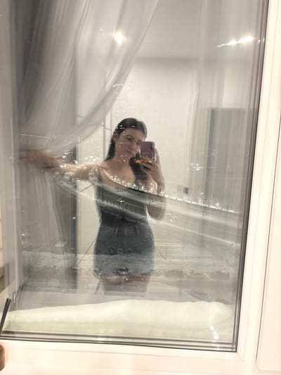 A woman in a form-fitting denim dress takes a selfie in front of a window. The window reflects a cityscape at night, superimposed onto her image. A white curtain is partially visible on the left side of the frame. The window's white frame surrounds the scene. The reflection creates a blurred and layered effect.