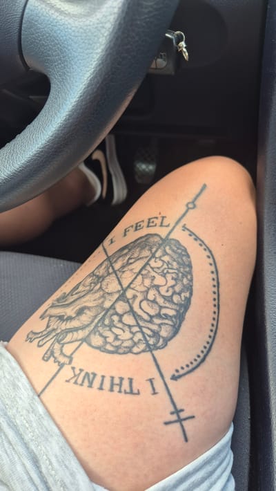 The image shows a close-up of a person's thigh with a tattoo of a brain. The tattoo is in black ink and depicts a stylized brain with lines and text. The text "I FEEL" is visible above the brain, and "I THINK I" is visible below. There are also some geometric lines and symbols around the brain. The person is wearing a grey t-shirt. The background shows parts of a car interior, including the steering wheel and a car key. There is no visible indication of the location being Kabile, Bulgaria.