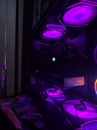 The image shows the interior of a computer case with multiple purple LED fans and lighting. The computer components visible include a graphics card labeled "INFINITY," a CPU cooler with a Cooler Master logo, and RAM modules labeled "DDR4 BEAST FURY." The computer is positioned near vertical blinds, suggesting it is indoors. On the left side of the image, there is a bottle with a purple liquid, and in the foreground, there are several packaged items, some of which appear to be food items or snacks. The lighting is predominantly purple, creating a moody and high-tech aesthetic. There are no people or any indication of activity. The scene is static. The location of Aleksandrovo, Bulgaria is not depicted or implied in the image.