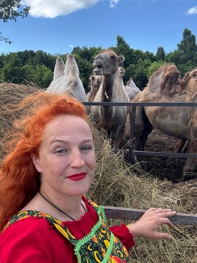 The image captures a woman with vibrant red hair and red lipstick taking a selfie outdoors. She is wearing a red dress with a colorful, embroidered bib and green trim. Behind her, several camels are visible in an enclosure. One camel in the foreground appears to be chewing or spitting. The enclosure is filled with hay, and a metal fence separates the woman from the animals. In the background, there is a line of lush green trees against a blue sky with scattered white clouds. The overall scene suggests an outdoor setting, possibly a farm or a zoo, in or near the city of Tri Ozera, Russia, given the context provided.