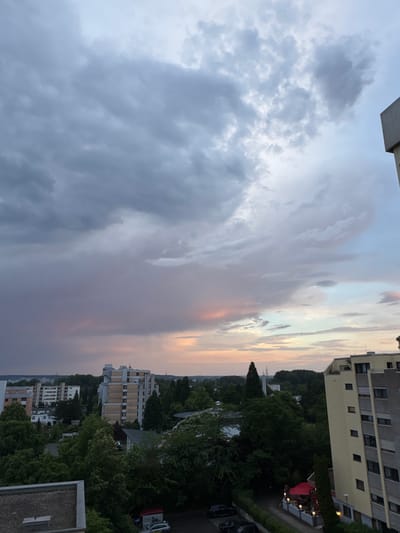 The image depicts a view of the city of Sindelfingen, Germany, at dusk. The sky is filled with dramatic clouds, showing a mix of dark gray, purplish, and pastel pink and orange hues, indicating an approaching storm or the aftermath of one. A few wispy clouds catch the fading sunlight.
Below the sky, the urban landscape unfolds. Several multi-story apartment buildings are visible, some with light-colored facades and dark-framed windows. Tall green trees populate the area between the buildings, creating a dense canopy in places. In the foreground, parts of two more buildings are visible, one with a flat, dark roof and another with a light-colored, textured surface.
A street is partially visible with parked cars. One car appears to be a police vehicle, marked with a red and white stripe and the word "Polizei" on its side. There is also a red umbrella and outdoor seating area with lights illuminated, suggesting a restaurant or cafe. Electrical pylons can be seen in the distance against the horizon, silhouetted by the dimming light. The overall impression is one of a residential area in a German city during twilight, under a brooding sky.