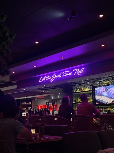 Here's a forensic description of the image:
**Location:** Appears to be a restaurant or bar interior in Boca Raton, Florida, United States, based on the presence of the establishment's name, "Let the Good Times Roll", which is a known establishment in that city.
**Environment:** Dimly lit indoor space. The dominant colors are purple (from the neon sign and ambient lighting) and darker tones (blacks, grays, dark browns). The ceiling has recessed lighting. The setting suggests a contemporary or upscale establishment.
**Objects:**
* A large neon sign reading "Let the Good Times Roll."
* Bar with a display of alcohol bottles.
* High-top bar chairs with a woven design.
* Dining tables with patrons seated at them.
* A screen showing a video.
**People:**
* Patrons sitting at the bar and tables.
* Details of faces are not clear due to the low light and focus.
**Actions/Interactions:**
* People appear to be dining and socializing.
* The scene represents a typical evening at an upscale restaurant or bar.
