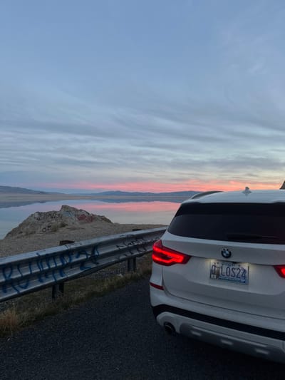 The rear of a white BMW SUV is positioned near a roadside barrier with graffiti, facing a stunning sunset. The license plate is visible, reading "0 LOS24" and indicating it is from Oregon. The landscape features a body of water reflecting the sunset, flanked by distant mountains. The sky displays streaks of pink, orange, and blue.