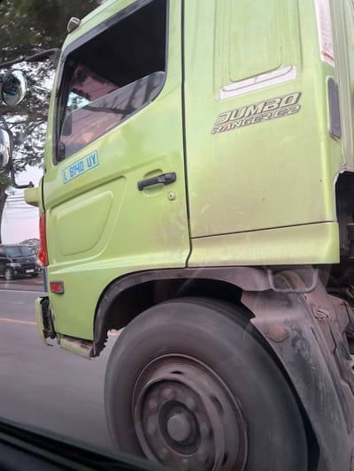 The scene depicts the passenger side of a light green Hino "JUMBO RANGER G2" truck in motion on a paved road in Jekulo, Indonesia. The truck's cabin features an open window, a closed door with a black handle, and a blue sticker reading "L 8140 UY" applied to the lower door panel. A side mirror assembly is partially visible on the far left. The large, dark-colored rear wheel of the truck displays significant motion blur, indicating forward movement. In the background, a dark-colored van or SUV is visible on the road, alongside out-of-focus green foliage and trees under a light sky. The image is captured from a slightly lower vantage point, likely from an adjacent moving vehicle. No individuals are visible.
