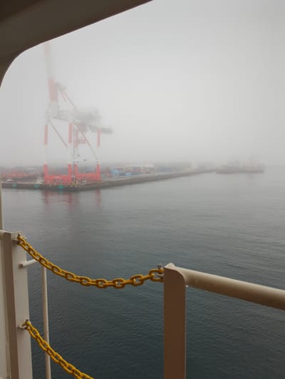 This is a photograph taken from a high vantage point, looking down at a foggy harbor scene in Tōkai, Japan. The primary subjects are large, red and white cranes, characteristic of a container port, partially obscured by thick fog. These cranes are positioned along a pier where shipping containers are stacked, suggesting an active cargo loading or unloading operation. Several ships are visible in the distance, also softened by the fog, indicating maritime traffic.

The overall scene is dominated by the ethereal quality of the fog, which creates a sense of mystery and limits visibility. The water of the harbor is a deep, textured gray-blue. In the foreground, a railing and yellow chains are visible, suggesting the photograph was taken from a ship or a structure overlooking the port.

The time of day is difficult to determine precisely due to the overcast conditions, but the diffused light suggests it is daytime. The weather is clearly foggy and likely cool. The location is implied by the presence of shipping cranes and containers, as well as the explicit mention of Tōkai, Japan. No people are clearly discernible due to the distance and fog.

The mood of the image is somewhat somber and atmospheric, due to the heavy fog and muted colors. There is no visible text in the image. The focus is on the industrial activity of the port juxtaposed with the natural atmospheric phenomenon of fog.