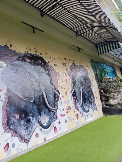 A mural of two elephants appears on a beige wall, seemingly bursting through a layer of bricks and earth. The left elephant is prominent with a detailed head and trunk. To the right, another elephant emerges similarly, though partially obscured. The wall is painted with fragmented debris, suggesting the impact of the elephants. Beneath the mural, bright green artificial turf provides a stark contrast. Above, a striped canopy and spotlights cover the area. To the right of the elephants, a faux rock formation is present.