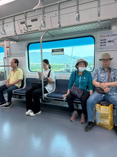 The image depicts the interior of a passenger train car, likely in Yangju, South Korea, with several individuals seated. The train car features blue seats, metal handrails, and windows offering a view of a mountainous landscape under a bright sky.
In the foreground, a man wearing a patterned blue and white short-sleeved shirt, blue jeans, and a straw hat is seated on the right. He has his arms crossed and appears to be looking forward. Next to him, a woman wearing a teal jacket, black pants, and a straw hat is seated. She has a white mask covering her mouth and nose, and her left arm is resting on her lap. She is looking to her left.
To the left of these two individuals, a woman wearing glasses, a white t-shirt, and black pants is seated, holding a smartphone and wearing earbuds. She is looking down at her phone. Further to the left, a man wearing a light yellow short-sleeved polo shirt and dark pants is seated, looking out the window.
Various objects are visible, including personal bags and a bright yellow tote bag with text that appears to be an environmental message. Korean text is visible on signs within the train car, indicating its location and amenities. The overall environment is clean and functional, consistent with public transportation.