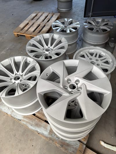 In Mol, Belgium, a collection of silver alloy car wheels are displayed on a concrete floor. Some wheels are stacked, while others are placed individually. A wooden pallet lies to the left of the scene, and another pallet with stacked wheels is visible in the background. The wheels appear to be newly finished or prepared for refurbishment, with a consistent silver finish. The environment suggests an industrial or workshop setting.