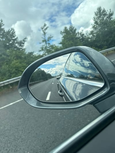 The image is a close-up, angled shot of a car's side-view mirror, reflecting a roadway scene. The mirror itself is dark grey, with visible scuff marks and a slight crack at the bottom.

In the reflection, a highway stretches into the distance, with a white line marking the right lane. Another vehicle, likely a car, is visible ahead in the reflection, though its details are blurred. The sky above is a mix of bright blue and fluffy white clouds, suggesting a partly cloudy day. Trees with green foliage line the side of the road, and a metal guardrail is visible to the right of the road in the reflection.

Outside the mirror, the blurred foreground shows trees and a bright, cloudy sky, indicating the general environment the car is traveling through. The perspective suggests the photo was taken from inside the car, looking towards the side mirror. There is no visible text in the image. The lighting suggests it is daytime, likely mid-morning or afternoon due to the brightness of the sky. The scene depicts travel on a highway, with the weather appearing fair.