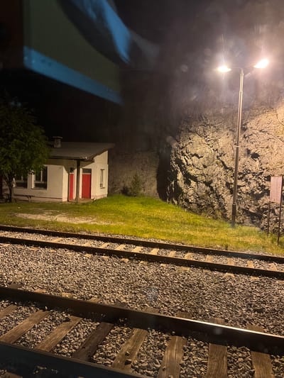 The image depicts a night scene alongside a railway track, likely in Lopate, Montenegro. The track is bordered by crushed gravel and wooden sleepers. Adjacent to the track is a strip of green grass. Beyond the grass, a small white building with red doors is visible. A rocky cliff face rises behind the building. A streetlight illuminates the scene. The left side of the image suggests the view is from inside a vehicle, due to the presence of a blurry reflective surface.