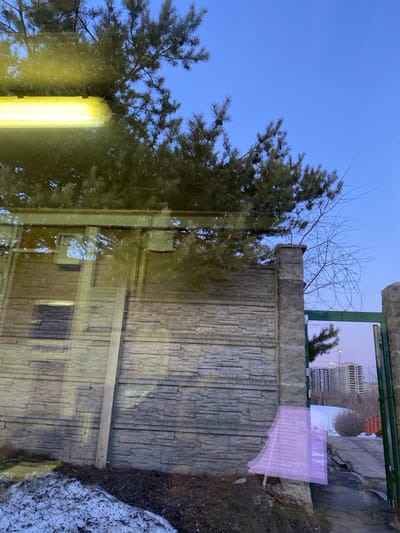 The image depicts an outdoor scene in Марфино, Russia, captured through a reflective surface, likely a window. The upper frame is dominated by a clear, pale blue sky and the dense green needles of a pine tree, with smaller bare tree branches visible to the right. A prominent, horizontal yellow-orange fluorescent light fixture is reflected across the upper-left quadrant.
The mid-ground features a textured, light gray concrete block wall, horizontally patterned to resemble stacked stone. To the right, an open dark green metal gate is visible, flanked by concrete pillars. Coiled barbed wire is present on top of the concrete pillar immediately to the left of the gate opening. Beyond the open gate, a dirt or unpaved road curves upwards, and in the distant background, multiple tall, modern multi-story buildings under construction, including a visible construction crane, stand against the sky.
The foreground shows dark earth with patches of melting snow. A vertical light-colored beam, potentially a wooden post, is positioned near the left side of the concrete wall. A translucent, angular magenta/purple shape is superimposed over the lower-right section of the wall and ground, consistent with a reflection or optical anomaly. No individuals are present in the scene.