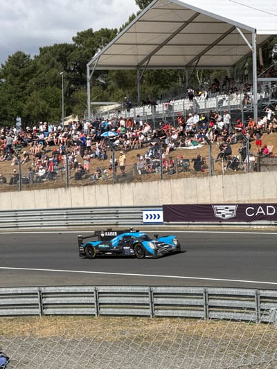 A Formula E race car, sponsored by Kaiser and displaying the number 25, speeds along a track. Spectators are seated in a large tented area beside the track, which is lined with chain-link fencing and crash barriers. A promotional banner with the Cadillac logo and a downwards-pointing arrow is also visible. The sky is overcast.