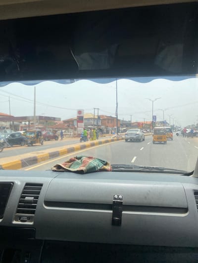 View from inside a vehicle traveling along a city street in Alakia, Nigeria. A light-colored sedan and a yellow tricycle taxi are visible, among other vehicles. People are walking alongside the road. Buildings and utility poles line the street. A fuel station is present. The dashboard of the vehicle is visible.