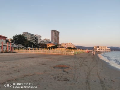 The image depicts a serene beach scene in Follonica, Italy, during what appears to be late afternoon or early evening. The foreground is dominated by a sandy beach with tire tracks from vehicles visible. In the middle ground, a white picket fence separates the beach from a row of beach umbrellas and lounge chairs, many of which are green and yellow. Behind the umbrellas, several buildings are visible, including a tall, modern high-rise and other multi-story structures that appear to be hotels or apartments. To the right, a white building with a pier-like structure extends over the water, and two individuals are walking on the beach near the water's edge. The sea is calm, with gentle waves lapping the shore, and the sky is a clear, pale blue, transitioning to a warmer hue near the horizon. The overall atmosphere is peaceful and tranquil.