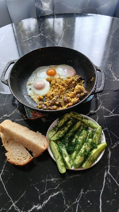 A bird's-eye view shows a rustic meal on a dark marble table, likely in Nadezhdenka, Russia. In a black cast-iron skillet, two fried eggs, one with a runny yolk, sit atop a bed of pilaf with chunks of meat. To the left, two slices of crusty white bread are stacked. To the right, a white plate holds sliced cucumbers generously sprinkled with fresh dill. The background is a dark, reflective surface with subtle marble patterns. The scene suggests a homemade, hearty meal.