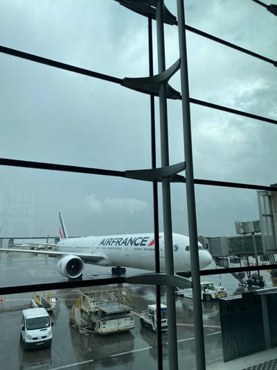 An Air France Boeing 777 aircraft is stationary on a wet tarmac at Charles de Gaulle Airport (CDG) in Mitry-Mory, France, viewed through large, multi-paned glass windows of an airport terminal. A grey passenger boarding bridge is connected to the aircraft's forward left door. Ground support equipment, including a white cargo loader and a white utility van, are positioned on the wet apron. The sky is heavily overcast, indicating dim, cloudy conditions, and the tarmac shows signs of recent or ongoing rain with visible puddles and reflections. No individuals are visible in the frame. The window structure consists of prominent grey vertical and horizontal beams with integrated supports.