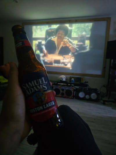 A partially visible person is holding a bottle of Samuel Adams Boston Lager in their right hand. The bottle is angled toward the camera, obscuring part of the view. In the background, a projection screen displays a scene from a movie or TV show, showing a man in a hat and suit. Speakers are stacked to the right of the screen. The image's details suggest an indoor setting. There is no evidence to establish the image was taken in Magdalena del Mar, Peru.