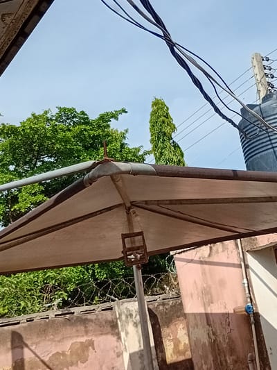 The image depicts an outdoor scene with a clear sky suggesting daytime with fair weather. The perspective is tilted, looking upwards. In the foreground, a light-colored awning is prominent, supported by metal frames. A grey gutter pipe runs along the edge of the awning, with a rusted connection visible.

Behind the awning, lush green trees fill a significant portion of the frame, their leaves vibrant and full. In the background, a building with a pinkish, weathered facade is visible. Electrical wires are strung across the sky, connected to an insulator on a pole, and a large cylindrical object, possibly a water tank, is also visible atop the structure. Barbed wire is strung across a fence in front of the trees, suggesting security measures. A blue water filter or valve is attached to a pipe on the building's exterior.

There is no visible text in the image. The scene appears to be in a residential or semi-urban area, likely in Kporva, Nigeria, given the context. No people are present, and the overall impression is one of everyday life with elements of infrastructure and nature. The tilted angle and close-up view of the awning and gutter might suggest the photo was taken from a window or balcony.