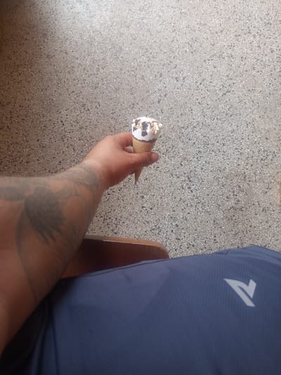 A first-person, top-down perspective photograph taken in Radishevo, Bulgaria, captures a seated individual holding an ice cream cone. The foreground features the person's right arm, which displays a large, dark, and faded tattoo resembling stylized wings or a predatory bird on the forearm, extending towards the bicep. The right hand firmly grips a waffle cone containing white or vanilla ice cream, generously topped with chocolate syrup drizzle and crushed nuts. To the left, a section of a dark brown, polished wooden armrest or chair edge is visible beneath the arm. Below the arm and to the left, the person's left thigh or lap is visible, covered by dark blue athletic fabric with a subtle mesh or pique texture. A white, stylized 'V' or arrow logo is prominently displayed on the fabric of the left thigh area. The background consists of a light grey aggregate concrete or terrazzo floor, composed of numerous small, dark grey and lighter flecks, appearing uniformly distributed and clean. The lighting suggests an indoor or covered outdoor setting, with brighter illumination towards the upper right quadrant of the floor.