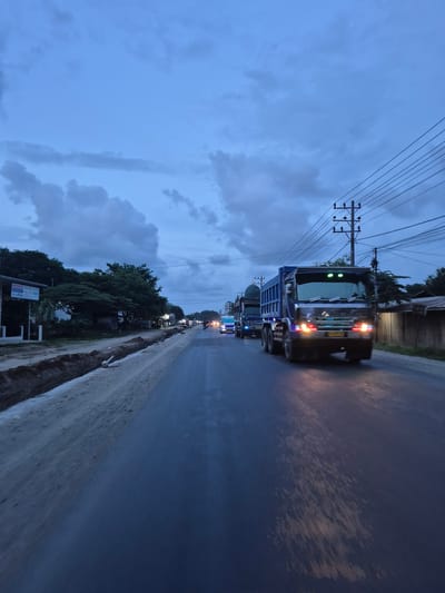A line of trucks travels along a road in Blangkeude, Indonesia, at dusk. Overhead power lines are visible above the road. Trees line the side of the road, and a ditch runs along the edge. The sky is cloudy.