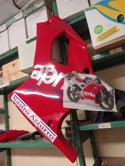 The scene depicts a red Aprilia motorcycle fairing suspended from a green metal storage shelf in what appears to be a storage room or workshop located in Fentange, Luxembourg. The fairing, likely a side panel, features "aprilia" in large silver lettering and "Champion World" in blue. A white sticker with "Nastro Azzurro" and "Birra chiara / Lager premium" in blue and red text is affixed to the lower left portion of the fairing. A color photograph of a red and silver Aprilia motorcycle is taped to the fairing's surface. The fairing is held by a thin green wire.
The surrounding environment consists of green metal shelving units against a light-colored wall. The top shelf holds several white cardboard boxes, some labeled "APRILIA." Below this, a white perforated plastic basket is visible, alongside a large cardboard box marked "CO2 Neutral" and "SELECTED QUALITY CLASS EXTRA." A lower shelf contains a folded red cloth, a black object, and a small wooden box. The shelf below that is labeled "Just_03." No individuals are present in the image.