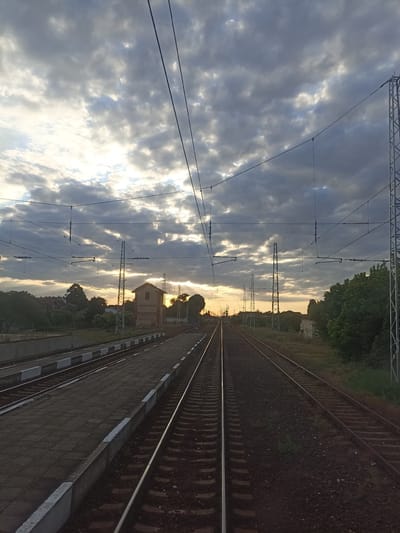 This image captures a view of a train station in Straldzha, Bulgaria, during what appears to be sunset or sunrise. The sky is filled with dramatic clouds, with streaks of sunlight breaking through, creating a moody atmosphere. In the foreground, multiple sets of train tracks stretch into the distance, bordered by a paved platform. The platform is marked with white and grey edges. Above the tracks, a complex network of overhead power lines and supporting structures is visible. To the left of the tracks, there's a low-lying building with a red roof and a tan facade, flanked by trees and houses. To the right, more trees and vegetation line the tracks. The overall scene is one of industrial infrastructure set against a natural, atmospheric sky.