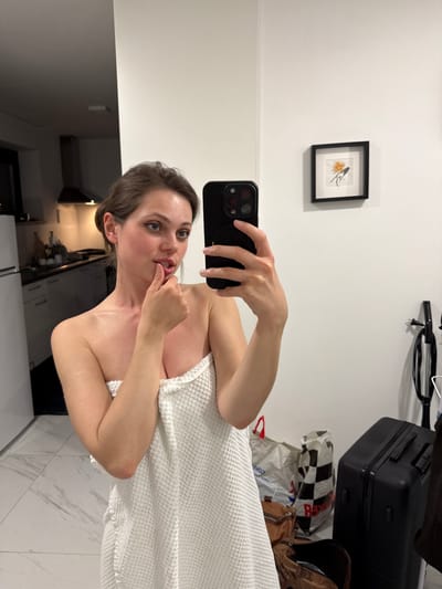 A woman wrapped in a white towel is taking a selfie in what appears to be an apartment. She has brown hair, fair skin, and is holding a black smartphone with a multi-lens camera. Her finger is near her mouth in a suggestive pose.
Behind her, the scene includes a white wall with a framed picture hanging on it, a suitcase and assorted bags/luggage. In the lower part of the image, one can see elements of a kitchen with cabinets and possibly a stove hood.
The location is not specified and nothing in the image indicates this is in Kranj, Slovenia. Therefore, no information exists to support that assertion.