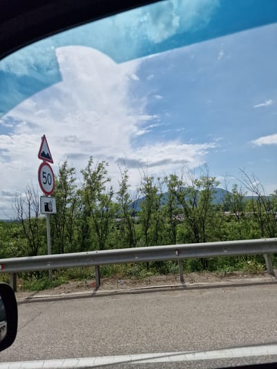 This is a photograph taken from inside a vehicle, looking out towards a rural roadside scene. The upper part of the image shows a blue sky with scattered white clouds, partially obscured by the vehicle's tinted windshield. Below the sky, a row of green trees with some bare branches forms a dense line. In front of the trees, a silver metal guardrail runs horizontally across the frame. To the left of the trees and slightly in front of them, a metal pole holds three road signs. The top sign is a red and white triangle with a black symbol depicting a bumpy road. Below it is a circular sign with a red border and a white background, displaying the number "50" in black. The bottom sign is rectangular with a white background and a black symbol of a camera with a flash. The ground in front of the guardrail is covered with dry grass and some scattered litter. A portion of the road surface is visible in the foreground, with white markings along the edge. A side mirror of the vehicle is visible in the lower left corner of the image. The photograph appears to have been taken during daylight, likely on a cloudy but bright day. The scene is consistent with a road leading into or out of a less developed area, potentially in a rural region of Russia like the vicinity of Змейка, which is known for its mountainous terrain and natural landscapes.