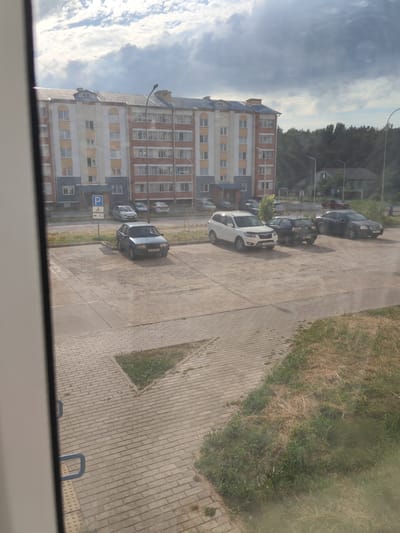 This image depicts a street scene outside an apartment building in Андрэеўшчынскі сельскі Савет, Belarus. The building is a multi-story structure with a pale yellow and orange facade, featuring numerous windows. In the foreground, several cars are parked in a lot. To the left, a dark blue sedan is parked, and further back, a white SUV, a black hatchback, and a black sedan are visible. A parking sign with a "P" and symbols for a bicycle and a wheelchair is also present. The ground is paved with concrete slabs and brickwork. Beyond the parking lot, there is a dense line of trees, and further in the distance, a few houses can be seen. The sky is partly cloudy with dramatic storm clouds and patches of blue sky, suggesting it might be late afternoon with the possibility of rain. The overall impression is a residential area on a typical day. There is visible text on the license plates of the cars.