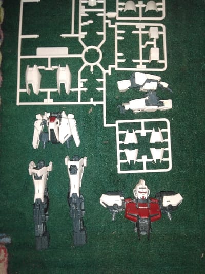 The image displays various plastic model kit components and partially assembled sections of a mechanical figure, consistent with a Gundam or similar mecha, laid out on a dark green, textured surface, likely felt or carpet. This scene is located in Quesada, Costa Rica.

Numerous white plastic parts remain attached to two distinct white sprues; a larger, irregular sprue occupies the upper and central portions of the frame, while a smaller rectangular sprue is situated in the middle-right. These sprues contain individual white armor panels and structural elements.

Several partially assembled sections of the model are also visible:
*   In the lower right quadrant, a complete head and upper torso unit is present, featuring a white head with red accents, and a main chest section in red and dark gray, flanked by white and dark gray shoulder assemblies.
*   In the lower-middle left, two distinct, partially assembled lower leg units are positioned vertically, primarily dark gray with white exterior armor panels.
*   In the middle-left, above the leg units, a partially assembled waist or hip section in white and dark gray is displayed horizontally.
*   In the middle-right, above the smaller sprue, two distinct, partially assembled arm units in white and dark gray are laid out horizontally.

No individuals are present in the image. No active assembly or interaction with the model parts is depicted.