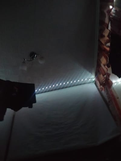 The image depicts a low-angle, indoor view of what appears to be a ceiling and adjacent wall. A light fixture with two globes is mounted on the ceiling, casting some light. A string of small, bright LED lights runs along the seam where the ceiling meets the wall, creating a glowing line. In the lower left quadrant of the image, a dark, indistinct object, possibly a piece of fabric or clothing, is suspended or draped. The wall to the right has a patterned fabric draped over it, with visible stripes of red, white, and possibly brown. The overall lighting is dim, with the LED strip and the light fixture providing the primary illumination. There are no visible people or any specific urban context that would allow for definitive placement within Beloozyorsky, Russia.