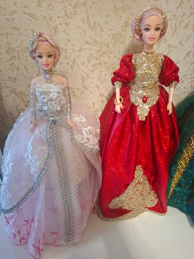 Two dolls are shown lying on a textured, cream-colored surface. The doll in the foreground is wearing a pink dress with white lace and silver trim. She has blonde hair with pink streaks and is wearing a silver headband and necklace. The second doll, behind the first, is wearing a sparkly red dress with gold embellishments and a red jewel on the chest. She has blonde hair with pink streaks and is wearing a gold headband. The dolls are posed as if standing, with their arms slightly bent. The background is a busy, textured fabric that appears to be lace or a similar material. The lighting is bright, suggesting an indoor setting with artificial light. There is no visible text in the image. The dolls appear to be decorative, possibly part of a collection or display.