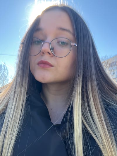 A close-up, front-facing outdoor portrait depicts a young adult female with long, straight hair, dark brown at the roots transitioning to blonde highlights. She wears round, light-framed glasses, and her lips are colored with reddish-pink lipstick. Her green eyes are open and directed towards the viewer. She is dressed in a black garment, visible around her neck and shoulders. The upper left background features a clear blue sky with a prominent, overexposed sun causing lens flare. Bare deciduous trees are faintly reflected in her glasses, and a light-colored multi-story residential building is partially visible in the right background. The scene is located in the city of Большевик, Russia.