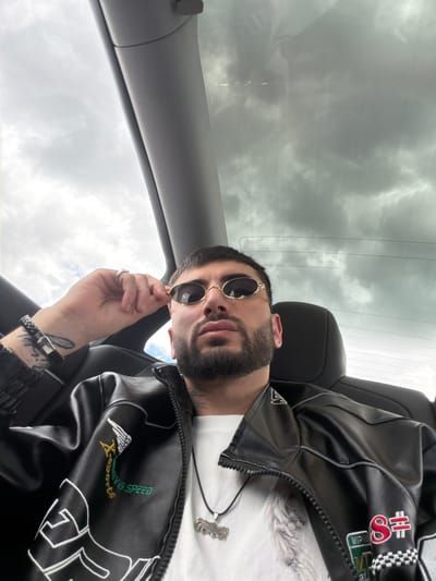 A man is sitting in a car, wearing sunglasses and a black leather jacket. The car is facing the sky, which is cloudy. It is not possible to determine the exact location in the city of Kotayk, Armenia from this image.