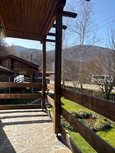 The image depicts an exterior scene in Liava Reka, Bulgaria, on a clear, sunny day, likely during early spring. The foreground features a covered veranda or balcony with a dark brown wooden plank ceiling. Its floor is paved with irregular, light-colored flagstones, exhibiting distinct shadow patterns. A sturdy wooden railing with square posts and three horizontal beams runs along the veranda's edge, partially obscuring the view of the immediate area below.
To the left, an adjacent traditional-style wooden building with a reddish-brown roof and a reflective window is visible. A dark wooden sign is affixed above its window. Beyond the railing, a well-maintained green lawn with a small, darker ground cover patch and a muddy depression is present. A secondary wooden fence borders this grassy area further out. Scattered deciduous trees, including a prominent birch, are bare of leaves. A stone-paved path extends from the left towards the right, past the adjacent building.
The background reveals a dirt or gravel road, where a white van or minibus is parked partially obscured by trees. Distant mountains, covered with sparse, bare trees, rise under a clear blue sky. Overhead utility lines and a street lamp are visible against the sky. No human subjects or direct interactions are depicted.