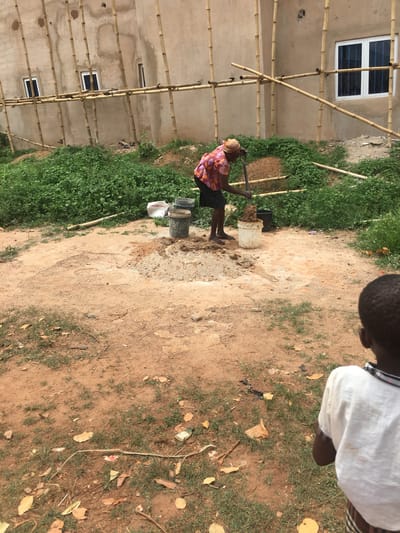 An outdoor scene in Isieke, Nigeria, shows a construction or labor site under natural daylight. In the center, a person with dark skin, wearing a colorful patterned short-sleeved shirt, dark shorts, and a head covering, stands barefoot on reddish-brown dirt. They are holding a long stick or tool, actively mixing what appears to be mud or concrete in a white plastic bucket. Another black bucket is visible nearby.
To the right, in the foreground, the back of a child's head and shoulder are visible, wearing a light-colored top, looking away from the camera.
In the background, to the left, is a light-colored building under construction or renovation. Numerous bamboo poles form scaffolding against its walls, a common construction method in the region. Several white-framed windows with dark panes are visible on the building.
The ground is a mix of reddish-brown earth and patches of green grass or weeds, with some scattered dry leaves and small pieces of litter. The bright lighting suggests it is daytime. No visible text is discernible in the image. The overall scene depicts manual labor associated with construction in a rural or developing setting.