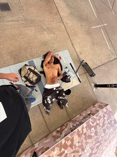 An overhead shot captures an urban outdoor scene in Bron, France, likely on a bright, sunny day. The primary subject is a young person with dreadlocks, lying shirtless on their back on a raised platform with gray and white tiles. They appear to be resting or sleeping, with one arm resting across their forehead and the other on their torso. They wear black pants with white graphic patterns and white boxer shorts, with blue and yellow sneakers visible on their feet.
Next to the person, a tan backpack, a clear plastic Evian water bottle, a mobile phone, and a black cap are placed on the tiled platform. Another individual, presumably the photographer, is partially visible in the foreground, showing their legs dressed in gray pants and blue sneakers, and their right hand. A belt with "POSCA" text is visible on the photographer's waist.
To the upper left of the main subject, a black scooter lies on the concrete ground. Further back, a black bollard or pole stands upright, displaying text including "UNABLE TO STOP," "NO STOP," "UNIVERSELLE," and "CAMPUS LYON 2," suggesting a university campus setting. To the right of the frame, a long, low structure is covered in an intricate mosaic of pink and brown tiles. The ground throughout the scene is rough concrete, featuring white painted lines and a metal grate drain.