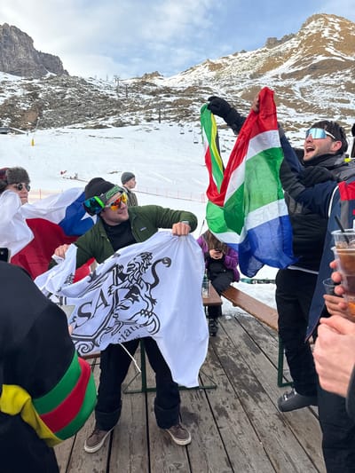 The image shows a vibrant outdoor gathering of people on a wooden deck at a ski resort, set against a backdrop of snow-dusted mountains and a ski slope in Alagna Valsesia, Italy. The weather appears clear and sunny.
In the upper part of the frame, a man wearing ski goggles on his head and a dark jacket is laughing heartily, holding a flag resembling the South African flag with its distinctive red, green, white, blue, and yellow colors. His hand, alongside others, holds plastic cups filled with beverages, indicating a festive toast or celebration. Below him, another man in a green jacket and ski goggles holds a large white flag emblazoned with a black crest featuring a rampant lion and partially visible text that reads "Valsesian". Another flag, red, white, and blue, is also visible near him.
Several other individuals are part of the group, dressed in winter clothing suitable for the mountain environment. Some are seated on a wooden bench, while others stand and socialize. Notable accessories include a fur-trimmed hat and a colorful red, green, and yellow striped beanie. A ski lift structure is discernible in the distant background, reinforcing the setting. The overall mood is jovial and celebratory, with people enjoying drinks and each other's company amidst the stunning alpine scenery.
